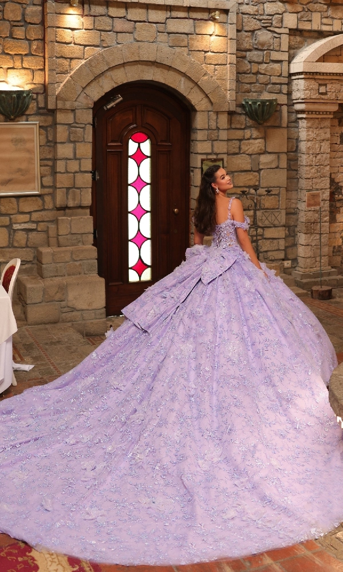 A woman in the off-shoulder Amarra 54243 quinceanera dress, featuring ornate lavender fabric with 3D floral appliques and an extended train, sits before a wooden door set in a stone wall.